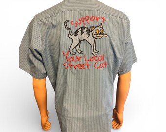 May include: Light blue short-sleeved button-up shirt with vertical pinstripes. The back of the shirt features an embroidered cartoon cat with the text "Support Your Local Street Cat" in red thread. The cat is gray and black.