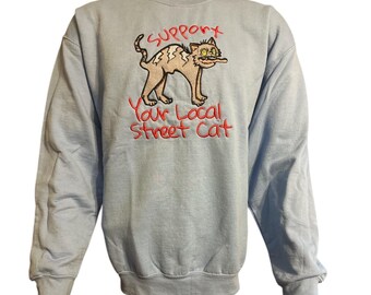 May include: Light blue sweatshirt with embroidered graphic of a cat holding a stick, with the text "Support Your Local Street Cat" in red. The cat is light brown with a white belly. The sweatshirt has long sleeves.