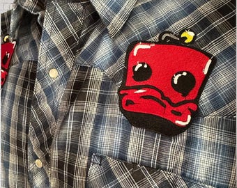 May include: Blue and black plaid button-up shirt with two red embroidered patches. The patches are cartoon-like with black outlines and details. The shirt has a classic collar and snap closures.