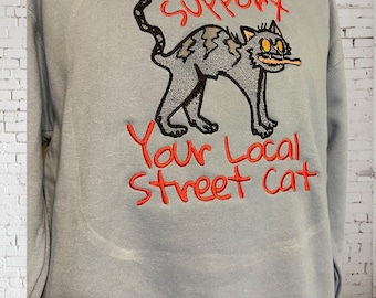 May include: A light blue sweatshirt with a cartoon cat graphic. The cat is gray with black outlines and yellow eyes. The text "Support Your Local Street Cat" is embroidered in red above and below the cat.