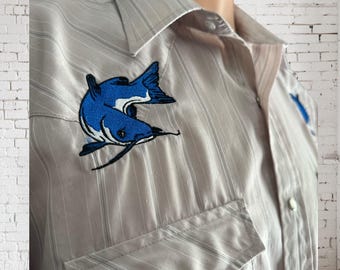 May include: A light gray striped button-up shirt with a blue embroidered catfish on the left side of the chest.
