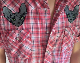 May include: Red plaid short-sleeve shirt with two front pockets and snap closures. Features two embroidered Sphynx cat patches with green eyes. The shirt has a classic western style.