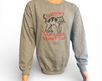 May include: A gray sweatshirt with long sleeves featuring an embroidered cartoon cat with the text "Support Your Local Street Cat" in red. The cat has a striped pattern and a stick in its mouth.