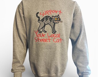 May include: A gray sweatshirt with a black cat graphic and the text "Support Your Local Street Cat" in red. The cat has yellow eyes and is holding a cigarette. The sweatshirt is a crew neck style.