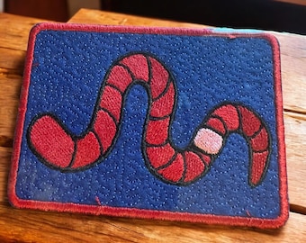 May include: Embroidered patch featuring a red worm design on a blue background. The worm has segmented sections and a black outline. The patch has a red border and is likely for clothing or accessories.