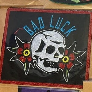 May include: Embroidered patch with a black background and a red border. The patch features a white skull with the words "BAD LUCK" in blue above it. Two red flowers with yellow centers flank the skull.