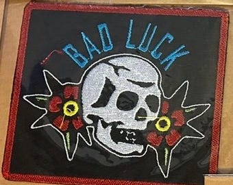 May include: Embroidered patch with a black background and a red border. The patch features a white skull with the words "BAD LUCK" in blue above it. Two red flowers with yellow centers flank the skull.