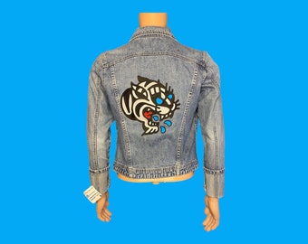 May include: A blue denim jacket with a black and white cartoon cat patch on the back. The cat has red and blue details and is dripping blue liquid.