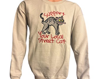 May include: A tan sweatshirt with a cartoon cat graphic and the text "Support Your Local Street Cat" in red. The cat is gray with yellow eyes and a pink tongue sticking out.