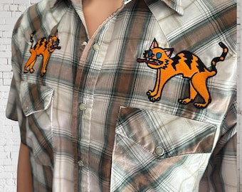May include: A plaid button-up shirt in shades of brown, green, and white. The shirt features two orange and black cat patches, one on each side of the chest. The shirt has two front pockets and a classic collar.