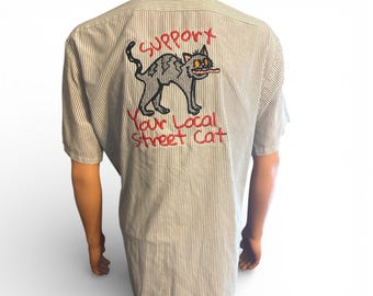 May include: Short-sleeved, striped shirt with a cat graphic on the back. The graphic features a gray cat with its tongue out, and the text "SUPPORT Your Local Street Cat" in red. The shirt has vertical stripes.