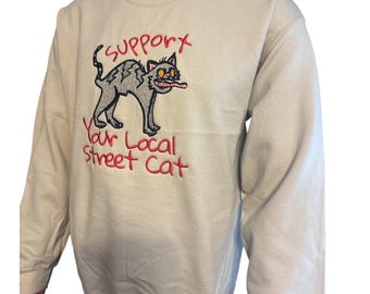 May include: A beige sweatshirt with a cartoon cat graphic. The cat is gray with orange eyes and a pink tongue sticking out. The text "SUPPORTx Your Local Street Cat" is embroidered in red above and below the cat.