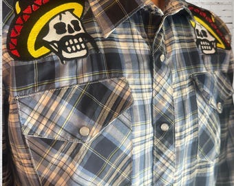 May include: Plaid button-up shirt in shades of blue, white, and yellow. Features two skull patches wearing sombreros on the shoulders. The shirt has two chest pockets and silver snap closures.