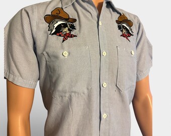 May include: A light blue and white striped short-sleeve shirt. The shirt features two embroidered patches of a raccoon wearing a cowboy hat and a red bandana. The shirt has two chest pockets and white buttons.