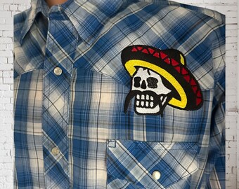 May include: Blue plaid button-up shirt with a skull patch wearing a sombrero. The patch is black, yellow, red, and white. The shirt has two front pockets and silver snap closures.