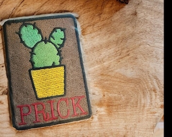 May include: A rectangular patch featuring a green cactus in a yellow pot, with the word "PRICK" in red letters. The patch has a brown background and a black border. The patch is placed on a wooden surface.