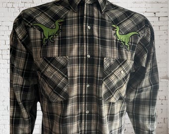 May include: A long-sleeved plaid shirt in black, white, and tan, featuring two green dinosaur patches on the chest. The shirt has a button-down front, two chest pockets, and a classic collar.