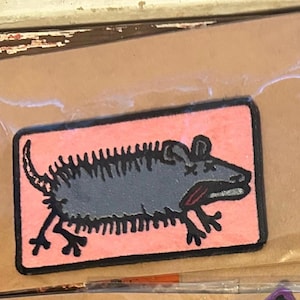 May include: A rectangular patch featuring a stylized gray rat with black outlines and details, set against a pink background. The rat has a long tail and is in a running pose. The patch is packaged in a clear plastic sleeve.