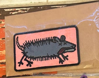 May include: A rectangular patch featuring a stylized gray rat with black outlines and details, set against a pink background. The rat has a long tail and is in a running pose. The patch is packaged in a clear plastic sleeve.