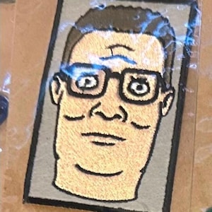 May include: Embroidered patch featuring a cartoon portrait of a man with glasses. The patch has a gray background and a black border. The man's face is tan, with brown hair and black glasses. The patch is encased in clear plastic.