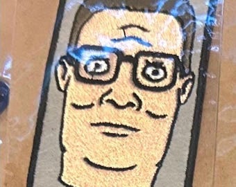 May include: Embroidered patch featuring a cartoon portrait of a man with glasses. The patch has a gray background and a black border. The man's face is tan, with brown hair and black glasses. The patch is encased in clear plastic.