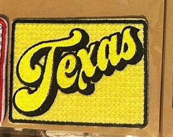 May include: A rectangular patch with the word "Texas" in yellow script with a black outline, set against a yellow textured background. The patch has a black border.