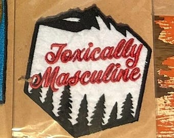 May include: A patch with the words "Toxically Masculine" in red script. The patch has a black mountain range at the top and black trees at the bottom, set against a white background.