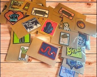 May include: A collection of embroidered patches on brown cardstock. Designs include a robot, a lizard, a cactus, a worm, and the text "Texas". Other patches feature a skull, a peach, and the text "Good Vibes".