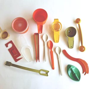 May include: A collection of vintage kitchen utensils in various colors, including orange, yellow, green, and white. The assortment features measuring cups, spoons, egg separators, and other cooking tools. The items are arranged on a white surface.