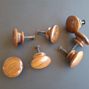 May include: A collection of seven wooden cabinet knobs in various shapes. The knobs are a warm brown color and have a glossy finish. Each knob has a metal screw for attachment. The knobs are arranged on a gray surface.
