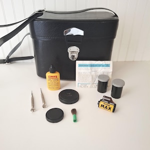 May include: A black camera bag with a silver clasp, accompanied by a Kodak lens cleaner bottle, film canisters, a film roll, and small tools. The kit includes a brush and a paper with text.