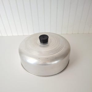 May include: A round, silver-colored aluminum lid with a smooth, metallic finish. The top surface features concentric circular ridges radiating from the center, topped with a small, black, cylindrical knob with a grooved, circular top design.