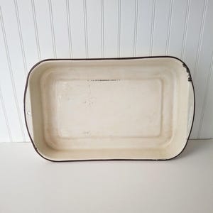 May include: A rectangular, off-white enamelware baking dish with a dark brown rim and handles. The vintage-style pan has a slightly distressed appearance, suggesting age and use. The interior is a creamy color, and the dish is empty.