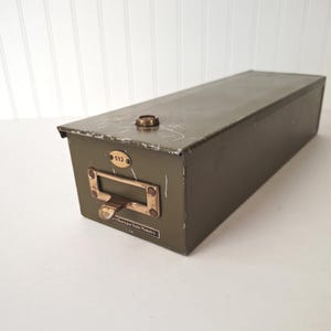 May include: An olive green metal drawer with a brass handle and label holder. The drawer has a small brass knob on top and a label reading "512". The drawer is rectangular and appears to be vintage.