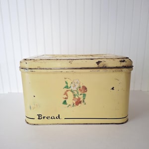 May include: Vintage yellow bread box with a floral design and the word "Bread" in black lettering. The rectangular box has a hinged lid and shows signs of wear, adding to its antique charm. The bread box is made of metal.