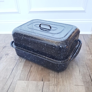 May include: A black enamel roasting pan with a lid. The pan has a rectangular shape with rounded corners and a speckled white pattern. The lid has a handle and a slightly domed top. The pan is sitting on a wooden surface.
