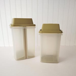 May include: Two vintage, clear plastic food storage containers with olive green lids. The containers are rectangular with a vertical divider in one. The lids are flat and slightly angled. The containers are on a white surface.