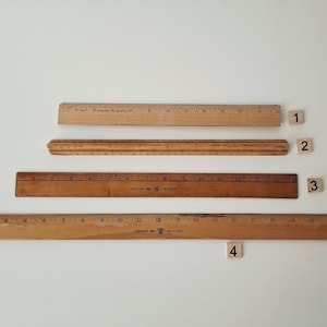 May include: Four wooden rulers of varying lengths are arranged on a white surface. The rulers are light and dark brown, with visible measurement markings. The longest ruler is approximately 24 inches long.