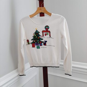 May include: A white sweater with a crew neck and long sleeves. The sweater features a colorful embroidered Christmas tree, presents, and ornaments design. The cuffs and hem are trimmed with black.