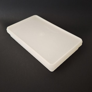 May include: A stack of off-white rectangular plastic food storage containers with lids. The containers are stacked on top of each other, with a slightly rounded edge. The background is black.