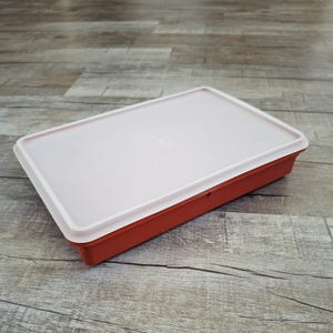 May include: A rectangular storage container with a deep terracotta orange base and a flat, semi-translucent off-white lid. The container features a simple, clean design with a small circular indentation centered on the top surface of the lid.