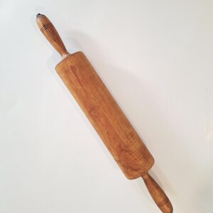 May include: A wooden rolling pin with a cylindrical barrel and two tapered handles on either end. The wood features a natural light brown grain pattern with visible texture and darker horizontal grooves carved into the handle sections.
