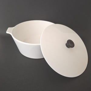 May include: A white ceramic serving bowl with a matching lid. The bowl has a rounded shape with a small handle on one side. The lid has a dark knob. The bowl and lid are on a dark gray background.