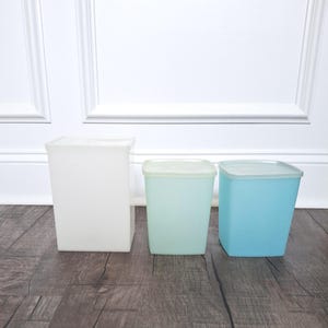 May include: Three square plastic containers in varying sizes and colors. The containers are white, light green, and blue, each with a matching lid. The containers are arranged on a dark wood-grain surface.