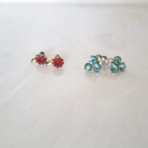 May include: Two pairs of earrings on a white surface. One pair features red, flower-shaped gemstones set in gold-colored metal. The other pair is a silver-toned ear cuff with blue and clear gemstones.