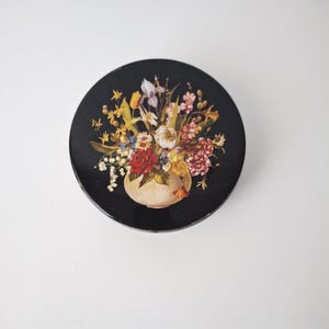 May include: A round, black decorative plate featuring a vibrant floral arrangement in a cream-colored vase. The bouquet includes red roses, yellow tulips, and various other colorful flowers. The plate has a glossy finish and is set against a white background.
