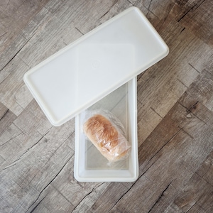 May include: A white rectangular food storage container with a clear lid, partially open. Inside, a loaf of bread is wrapped in clear plastic. The container is on a wooden surface.