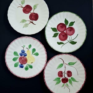 May include: Four white ceramic plates with scalloped edges, each decorated with hand-painted fruit designs. The fruits include apples, cherries, and a pear, with green leaves and red, green, and purple accents. The plates have a vintage aesthetic.