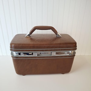 May include: A vintage brown Samsonite train case with a top handle and silver-toned hardware. The rectangular case has a textured surface, a brown handle, and two latches. The Samsonite logo is visible on the front.