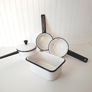 May include: A collection of white enamel kitchenware with black rims, including a rectangular baking dish, two round saucepans with long black handles, and one round saucepan with a matching lid and a black handle, all featuring a smooth finish.